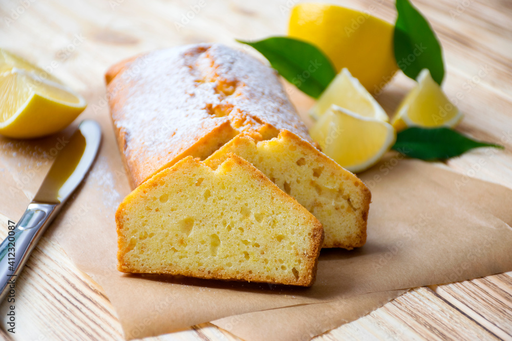 Loaf of traditional lemon pound cake with pieces of lemon, green leaves and knife on rustic wooden background. Slice of citrus pie by classic recipe. Homemade bakery, vegan dessert.