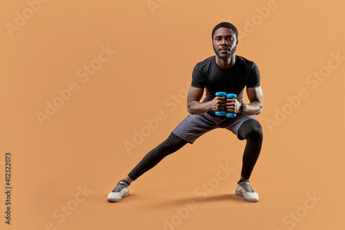 athletic man lunges with dumbbells in hands, do sportive exercises, lead healthy lifestyle, look at camera, isolated on brown background