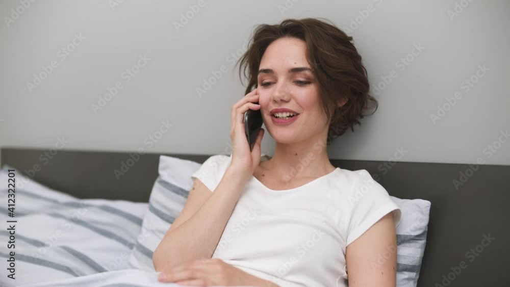 Happy young woman wear casual white t-shirt lying in bed rest relax spend time in bedroom lounge home in own room house hold in hands using mobile cell phone talking order delivery Real estate concept Happy young woman wear casual white t-shirt lying in bed rest relax spend time in bedroom lounge home in own room house hold in hands using mobile cell phone talking order delivery Real estate concept