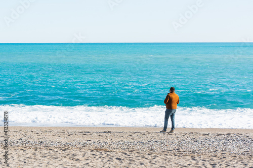 Lonely man stands with his back in a warm orange jacket stands on the beach and looking at horizon by the sea in winter in sunny weather. No face. Copy space for text