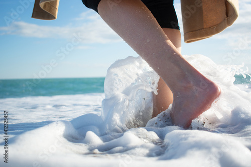 Beautiful legs of young girl, woman who goes towards sea, ocean in splashing sea foam against blurry background of blue sea and sky with white clouds in winter. Wave breaks against the girl's legs