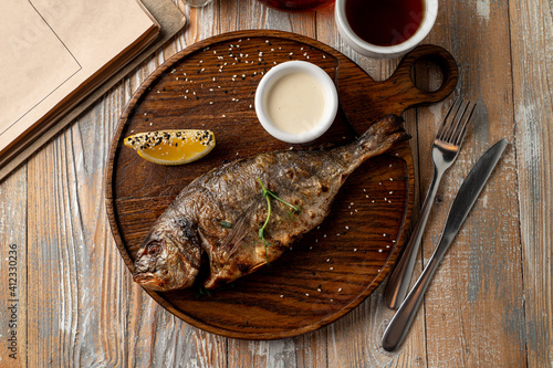 Whole grilled freshwater fish with head, garnished with sour cream and lemon, top view of rustic dinner, wooden background photo