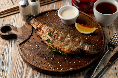 Whole grilled freshwater fish with head, garnished with sour cream and lemon, top view of rustic dinner, wooden background photo