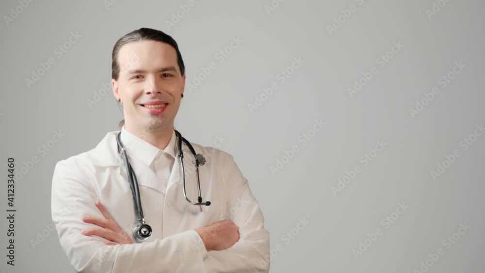 A male doctor with long hair walks, stops, smiles, and looks at the camera. Gray background. A male doctor with long hair walks, stops, smiles, and looks at the camera. Gray background.