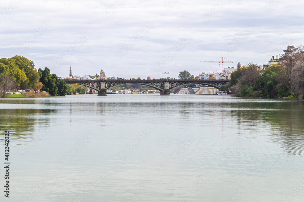 Urban landscape with the Guadalquivir river crossing the city of Seville in a cloudy day (Andalusia, Spain). Views of the Isabel II bridge, also known as the Triana bridge.