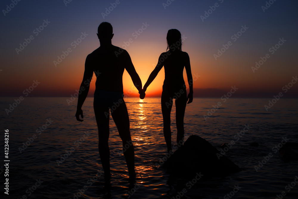 Vacation couple walking on sunset beach together in love holding hands. Happy young couple silhouette on sunset.