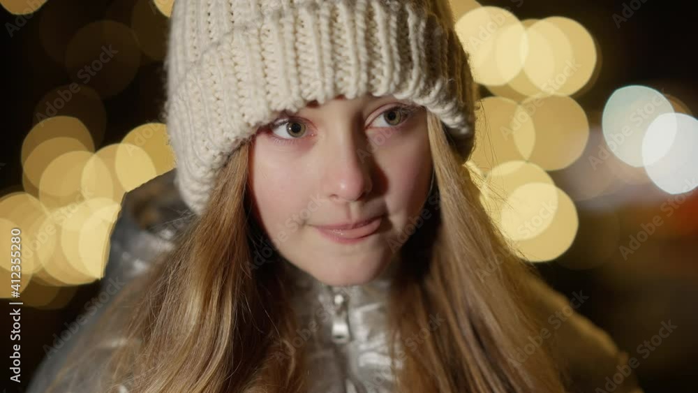 Headshot of charming brunette girl with hazel eyes standing at New Year night outdoors looking around. Close-up of cute Caucasian kid in dusk with Christmas lights at background. Headshot of charming brunette girl with hazel eyes standing at New Year night outdoors looking around. Close-up of cute Caucasian kid in dusk with Christmas lights at background.