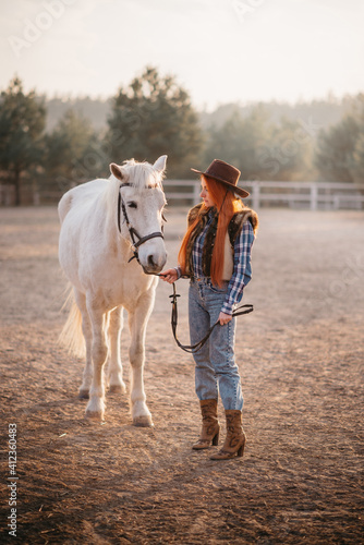 A young cowgirl woman stroking a horse in the sunset at her ranch.