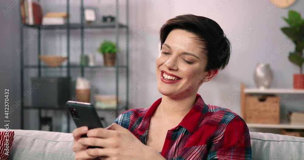 Smiling girl relaxing and sitting on sofa at home while using cell phone. Woman texting and chatting with friend, happy woman on couch hold smartphone and laughing while using mobile application Smiling girl relaxing and sitting on sofa at home while using cell phone. Woman texting and chatting with friend, happy woman on couch hold smartphone and laughing while using mobile application