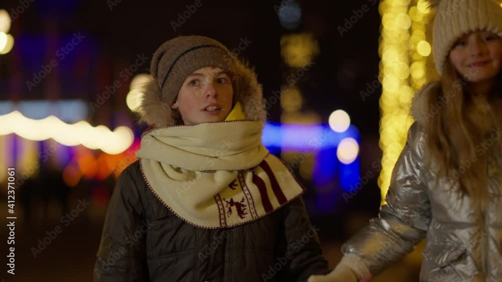 Positive cute boy and girl walking in slow motion with flashing Christmas lights at background. Positive Caucasian brother and sister or friends celebrating New Year in city outdoors. Positive cute boy and girl walking in slow motion with flashing Christmas lights at background. Positive Caucasian brother and sister or friends celebrating New Year in city outdoors.