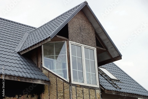 gray wooden attic of a private house with a large white window under a black tiled roof against the sky