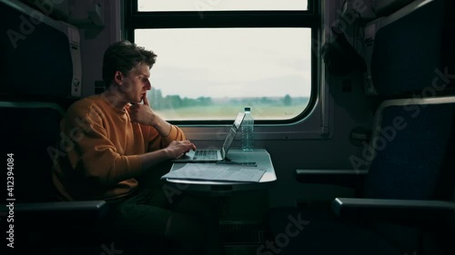 Handsome young man rides in the evening in a train compartment at a table with a laptop and takes photos from the window on a smartphone camera.Guy uses Internet on gadgets while traveling by train.