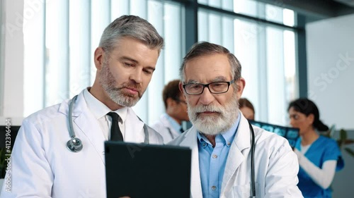 Healthcare and modern medicine. Serious senior doctor looking together with his colleague at the survey results showing at the digital tablet while preparing for serious treatment at the clinic photo