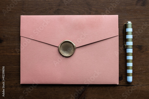 A salmon coloured envelope holding a wedding invitation with a gold wax seal, next to a small gold and white pen on a dark wooden desk, Toronto, Ontario, Canada.