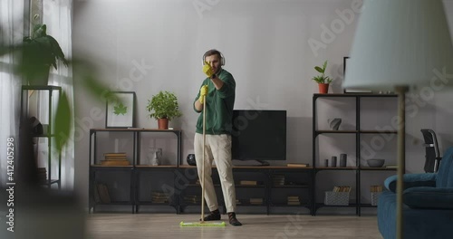 comical man is singing into mop during clean-up in flat, listening to music in headphones, imaging himself as superstar