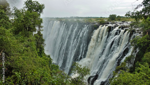 The powerful streams of Victoria Falls plunge into the abyss. The rocky channel of the Zambezi River is visible. In the foreground, green tropical vegetation. Zambia