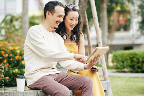 Happy handsome young Chinese man showing new application on tablet computer to girlfriend when they are sitting on bench in park