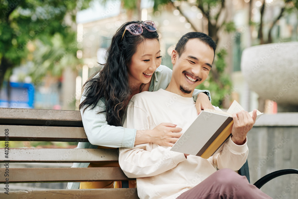 Pretty smiling young Asian woman looking in book of boyfriend reading book when sitting on park bench