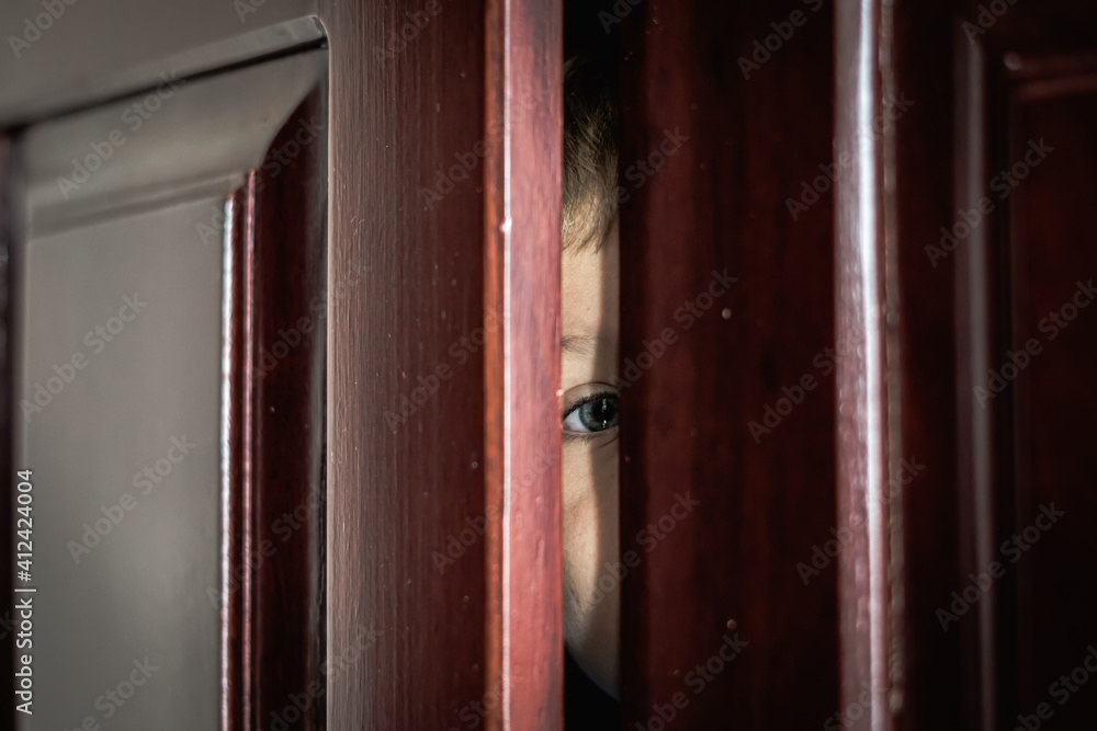 Obraz premium Sad boy hide in closet, close-up view of blue eye in ajar door slit home wardrobe. Child emotions. Warning symptom sign, treatment help for childhood adolescent depression illness, fears, family life