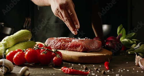 Chef sprinkling raw meat with salt on a background with vegetables. Cooker preparing meat in professional kitchen.