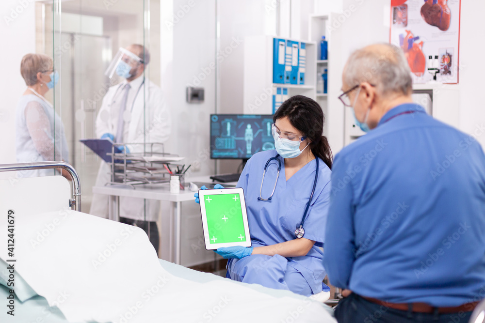 Patient looking at tablet with green screen during consultation in hospital room with nurse wearing face mask against coronavirus as safety precaution. Medical examination for infections, disease . Patient looking at tablet with green screen during consultation in hospital room with nurse wearing face mask against coronavirus as safety precaution. Medical examination for infections, disease .