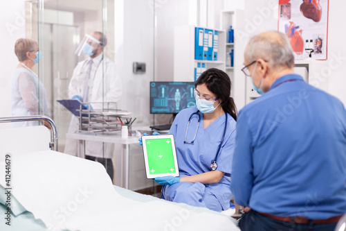 Patient looking at tablet with green screen during consultation in hospital room with nurse wearing face mask against coronavirus as safety precaution. Medical examination for infections  disease .