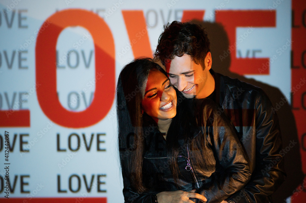 Intimate loving Indian and Caucasian couple with black jacket embracing. Lovers smiling in dark studio illuminated by light of projector with word love. Intimate loving Indian and Caucasian couple with black jacket embracing. Lovers smiling in dark studio illuminated by light of projector with word love.
