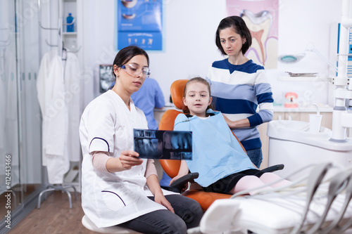 Dentistiry specialist talking about cavity procedure to kid wearint dental bib. Stomatologist explaining teeth diagnosis to mother of child in health clinic holding x-ray. photo