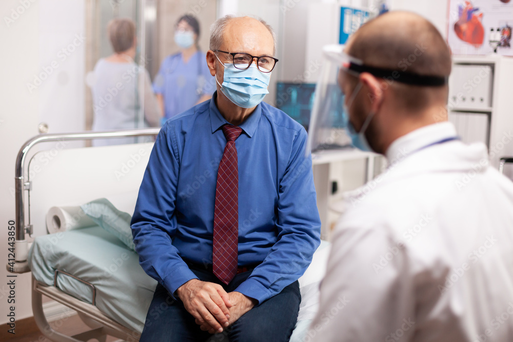 Obraz premium Doctor in the course of covid outbreak consulting senior man in examination room. Physician practitioner in the course of private modern clinic appointment during coronavirus pandemic.
