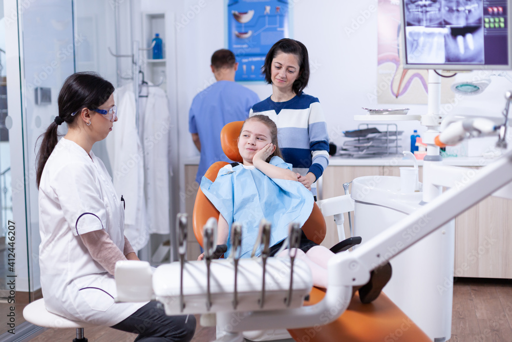 Obraz premium Little girl in pediatric dentist office touching face with painful expression because of tooth health issues. Child with her mother during teeth check up with stomatolog sitting on chair.