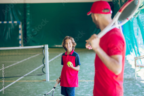 Young bearded man in a red cap holding a tennis racket and talking to a boy