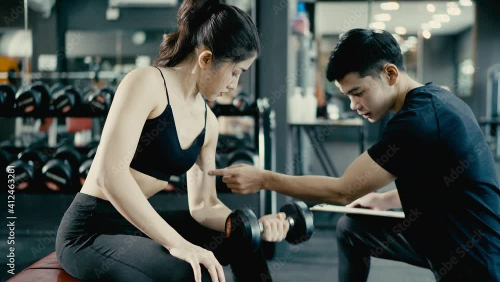Asian Slim girl exercising at fitness center. Young handsome trainer assisting pretty woman lifting dumbbells at gym. Asian Slim girl exercising at fitness center. Young handsome trainer assisting pretty woman lifting dumbbells at gym.