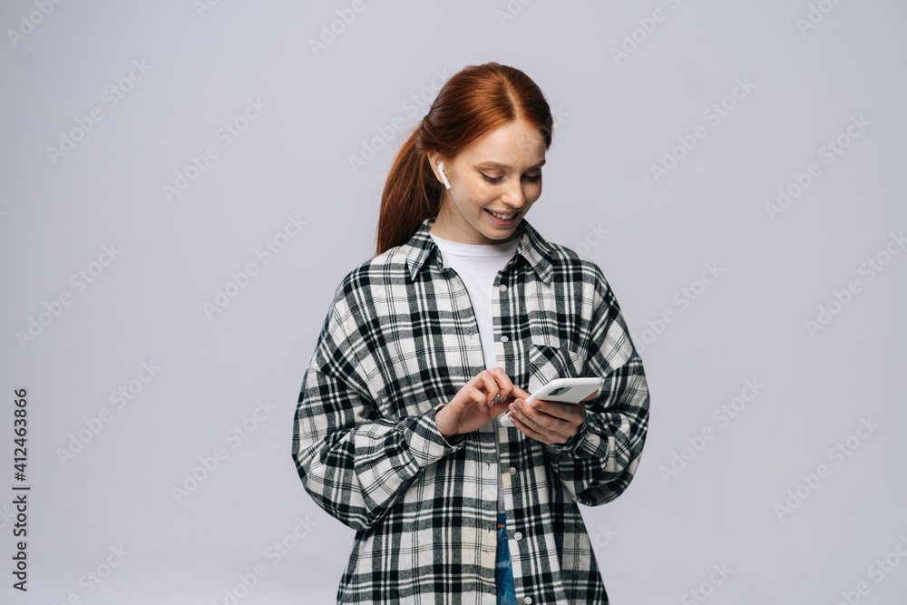 Photo of cheerful redhead young woman using modern technology headphones ears listen popular song choosing telephone next audio track against isolated white background. Photo of cheerful redhead young woman using modern technology headphones ears listen popular song choosing telephone next audio track against isolated white background.