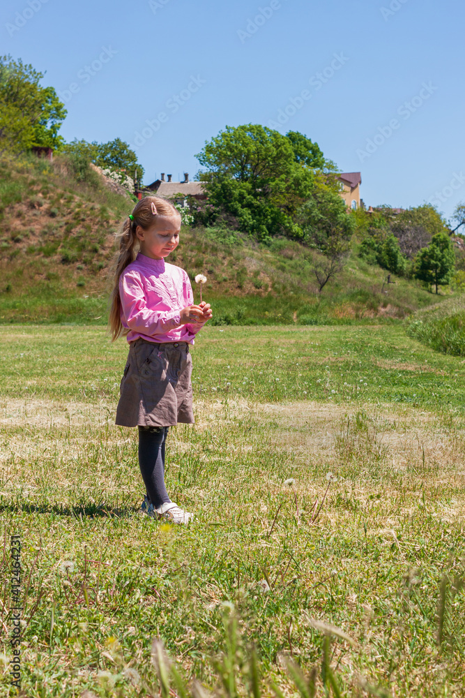 Positive emotional young lady with long blond hair smiles, looks on dandelion flower that holds in hand. Cheerful pretty child girl in skirt, pink shirt, stands on nature on sunny day. Happy childhood Positive emotional young lady with long blond hair smiles, looks on dandelion flower that holds in hand. Cheerful pretty child girl in skirt, pink shirt, stands on nature on sunny day. Happy childhood