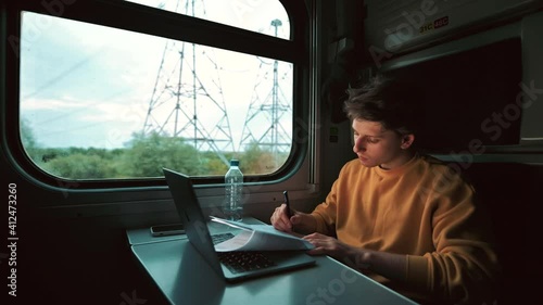 Wallpaper Mural Young man sits at a table in a train with a laptop and performs tasks on a piece of paper looking at a computer screen and writes with a pen. Torontodigital.ca