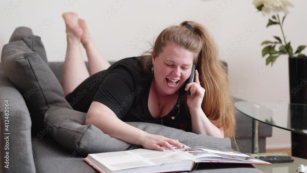 plump woman laughing happy while talking on the telephone and looking on a photo album lying on the couch plump woman laughing happy while talking on the telephone and looking on a photo album lying on the couch