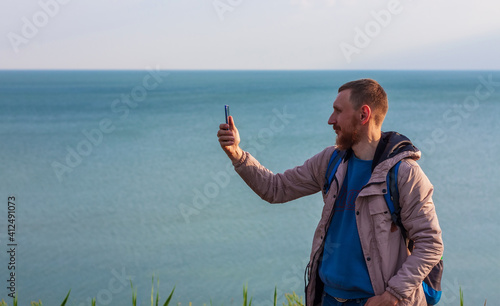 Bearded man in raincoat with backpack on sea landscape with blue cloudy sky background with smartphone in hand taking picture. Guy photographing nature on cell phone, active lifestyle travel concept.