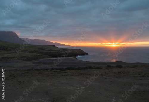 Beautiful sunset on the rocky atlantic coast in north west part of Gran Canaria island. Red orange sun with rays going down to the ocean. Dark blue clouds and cliffs in background.