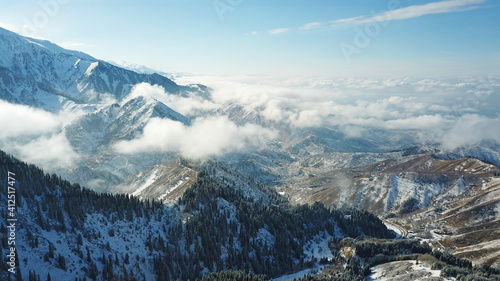 Snow forest in the mountains, above the clouds. View from above, from the drone. Coniferous trees are covered with snow. Clouds float along the gorge. Snowy hills and Sunny day. Tourists in mountains