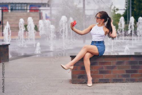 A woman in sunglasses sits and takes a selfie by the city fountain.