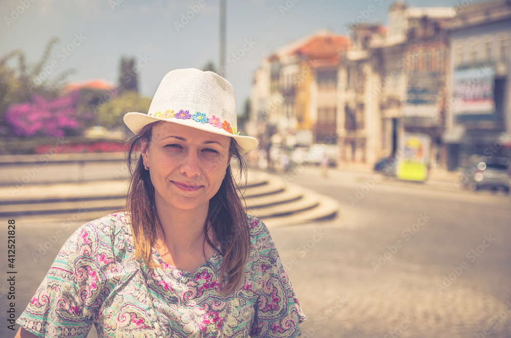 Young beautiful woman traveler with hat looking at camera posing and smiling in streets of Aveiro city historical centre in Portugal in sunny summer day on vacation, old houses buildings background