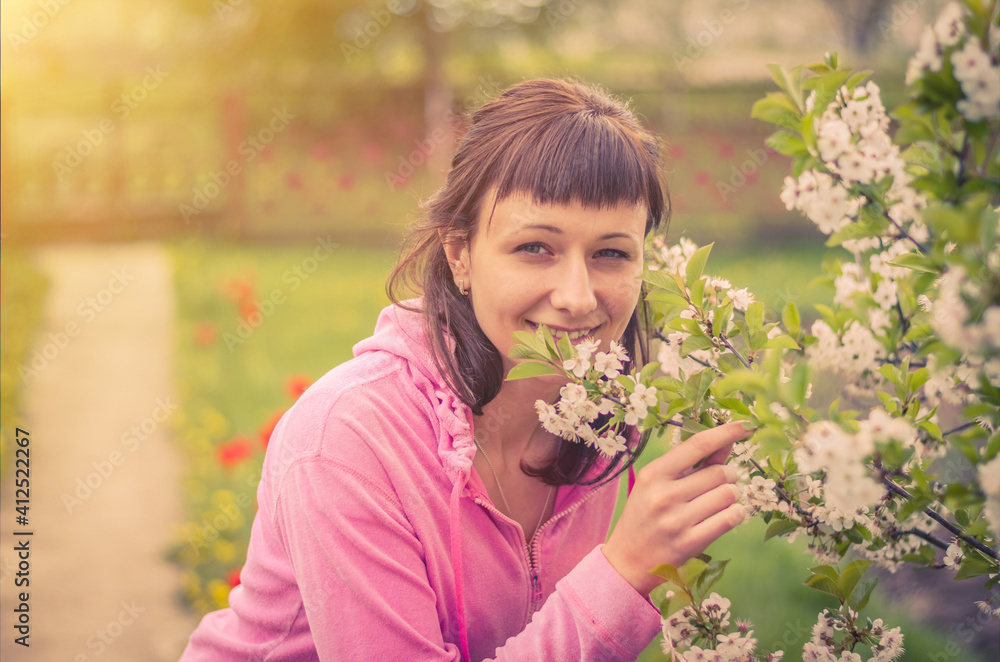 Young beautiful female woman looking at camera, smiling and smelling flowers of cherry tree blossom in spring sunny day with sun flare, close-up view, blurring garden and fence background Young beautiful female woman looking at camera, smiling and smelling flowers of cherry tree blossom in spring sunny day with sun flare, close-up view, blurring garden and fence background