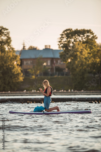 Fototapeta Naklejka Na Ścianę i Meble -  A young blonde woman on the phone on a SUP stand up paddle board in the river in summer, Golden hour