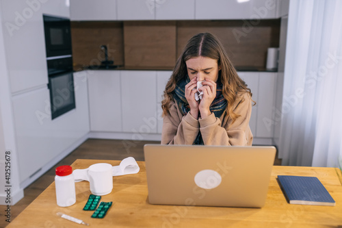 Tablou pe pânză Allergic woman blows her nose in a napkin, sits at a home office table, studying on a laptop, a sick girl got the flu, caught a cold, sneezes in tissue, has allergy symptoms, coughs, holding a napkin