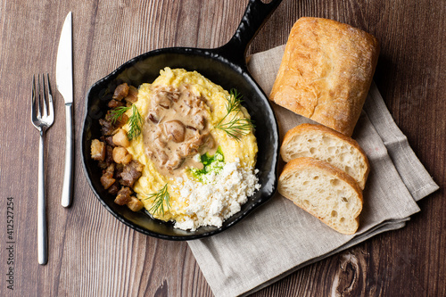 Banosh - a ukrainian dish made of cornmeal. Served in black pan with cracklings, mushrooms and hutsul cheese. Close-up isolated on a wooden background with cutlery, sliced bread and grey linen napkin. photo