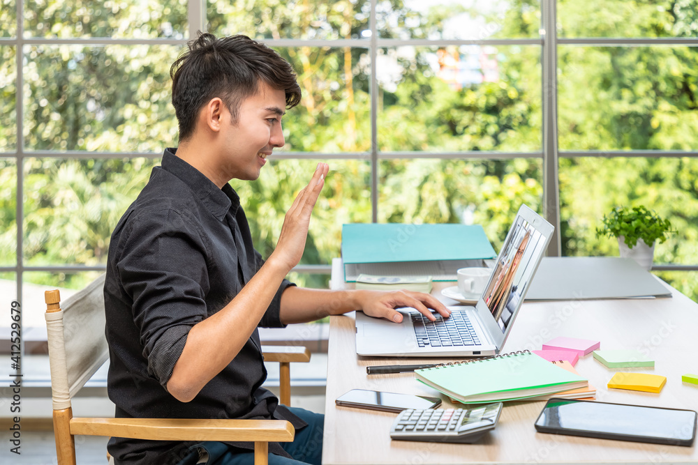 Young Asian businessman working at home with laptop in dining room, online meeting conference with colleague, and waving hand. Young Asian businessman working at home with laptop in dining room, online meeting conference with colleague, and waving hand.