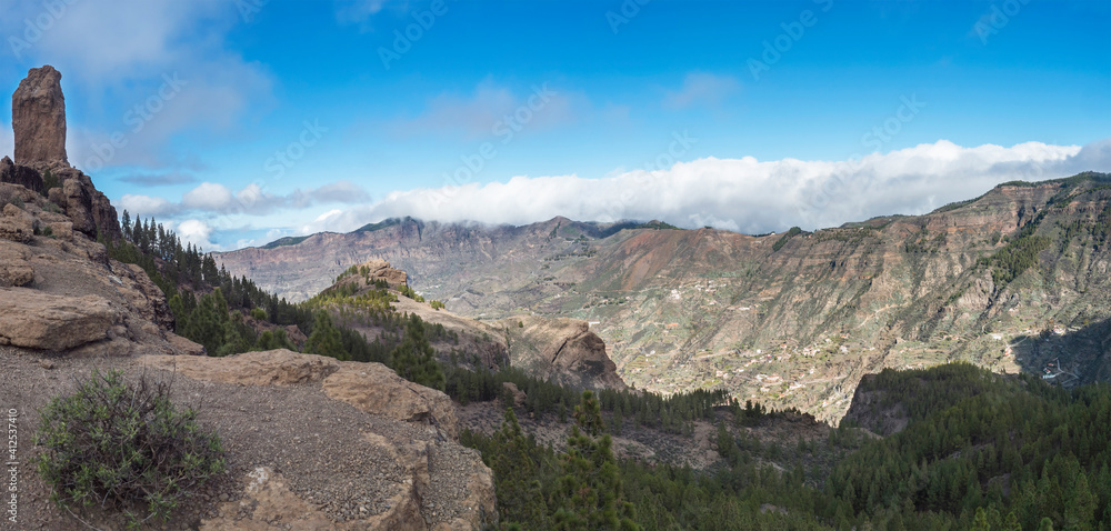 Panoramic view of Roque Nublo rock formation in inland central mountains from famoust Gran Canaria hiking trail. Green pine trees and blue sky background. Canary Islands, Spain.