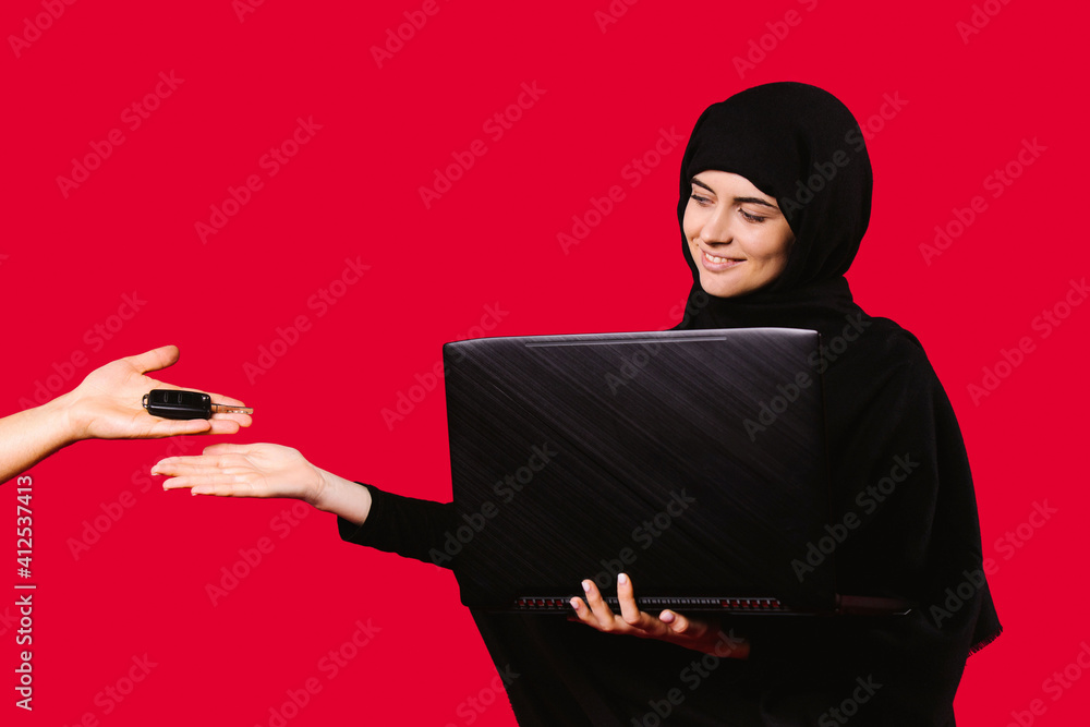A Muslim girl in a black hijab with a laptop in her hands with a smile receives the car keys from male hands. Red background. A Muslim girl in a black hijab with a laptop in her hands with a smile receives the car keys from male hands. Red background.