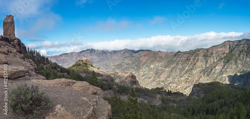 Panoramic view of Roque Nublo rock formation in inland central mountains from famoust Gran Canaria hiking trail. Green pine trees and blue sky background. Canary Islands, Spain.