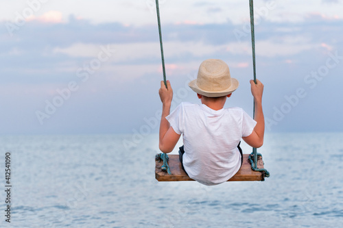 Teen in straw hat riding on rope swing over the water. Swing against the sky and the sea